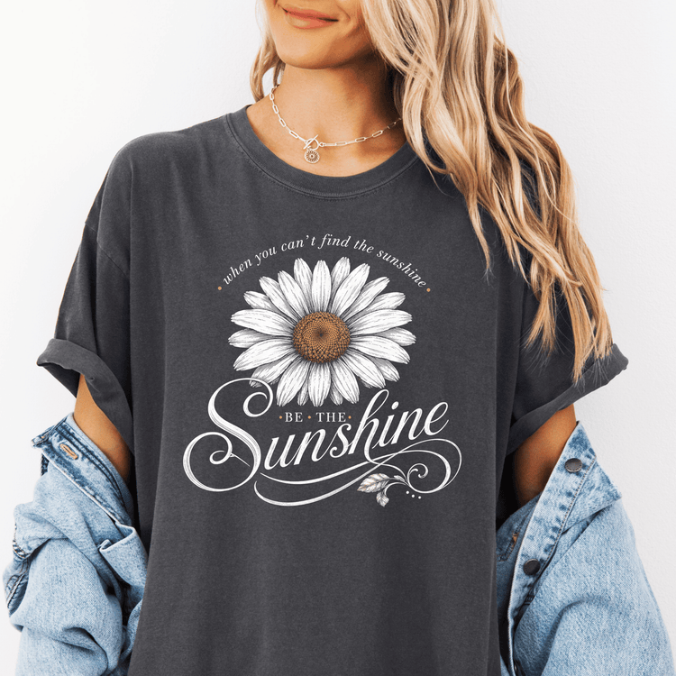 A woman wearing a gray t-shirt with a white daisy flower design and the text "When you can't find the sunshine, be the sunshine" printed on it.