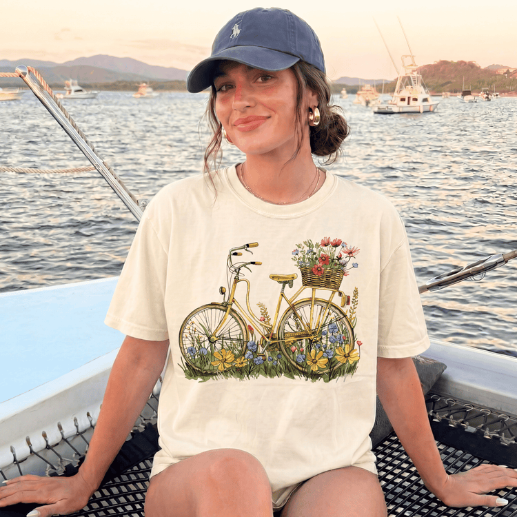 A woman wearing a white t-shirt with a bicycle graphic and flowers on it, sitting on a boat with a scenic background of water and boats.
