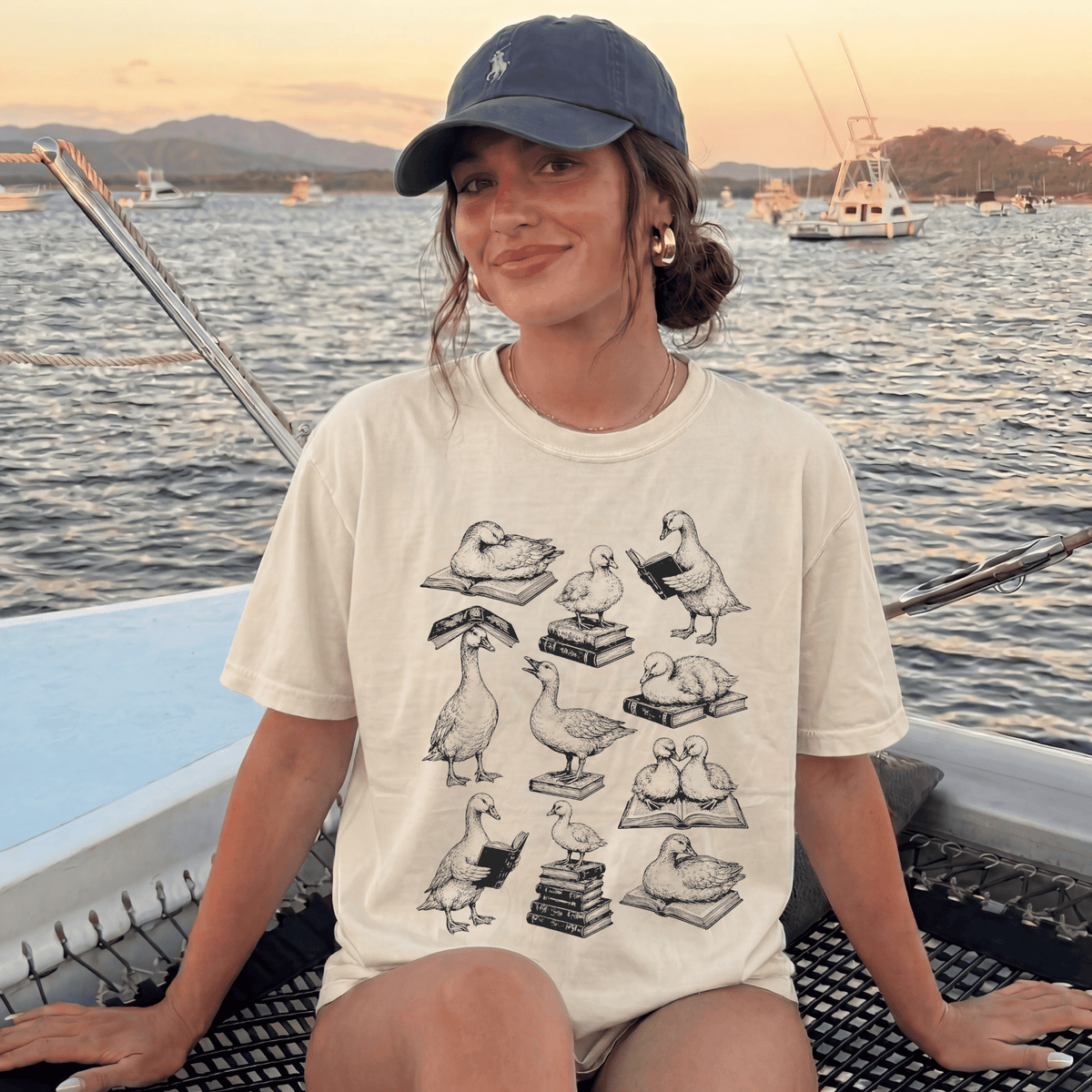 A woman wearing a white t-shirt with a graphic of ducks reading books while sitting on a boat in a body of water.