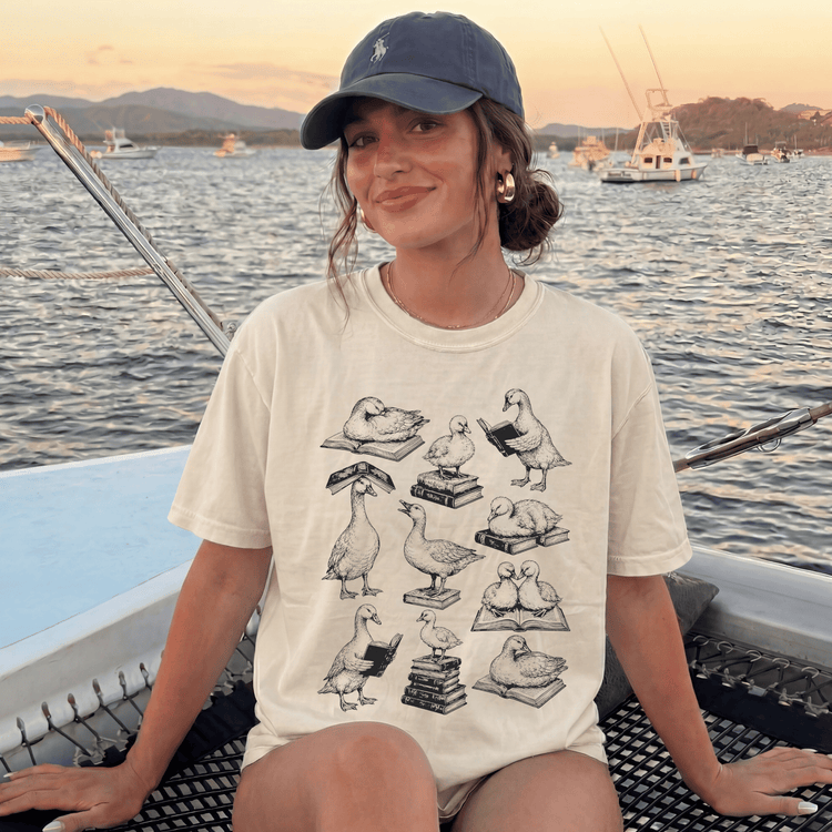 A woman wearing a white t-shirt with a graphic of ducks reading books while sitting on a boat in a body of water.