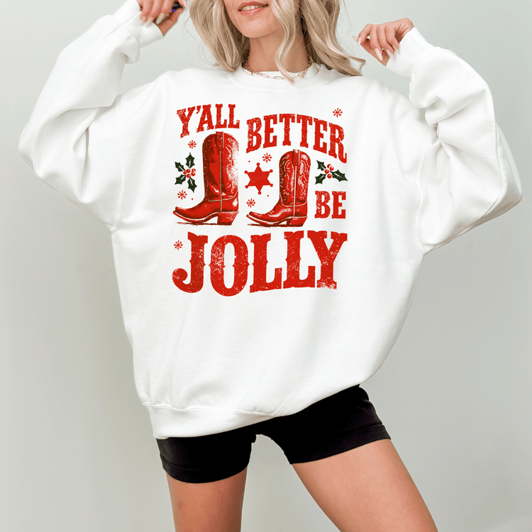 A woman wearing a white sweatshirt with a red graphic design that says "Y'all better be jolly" and features red cowboy boots and holly leaves.