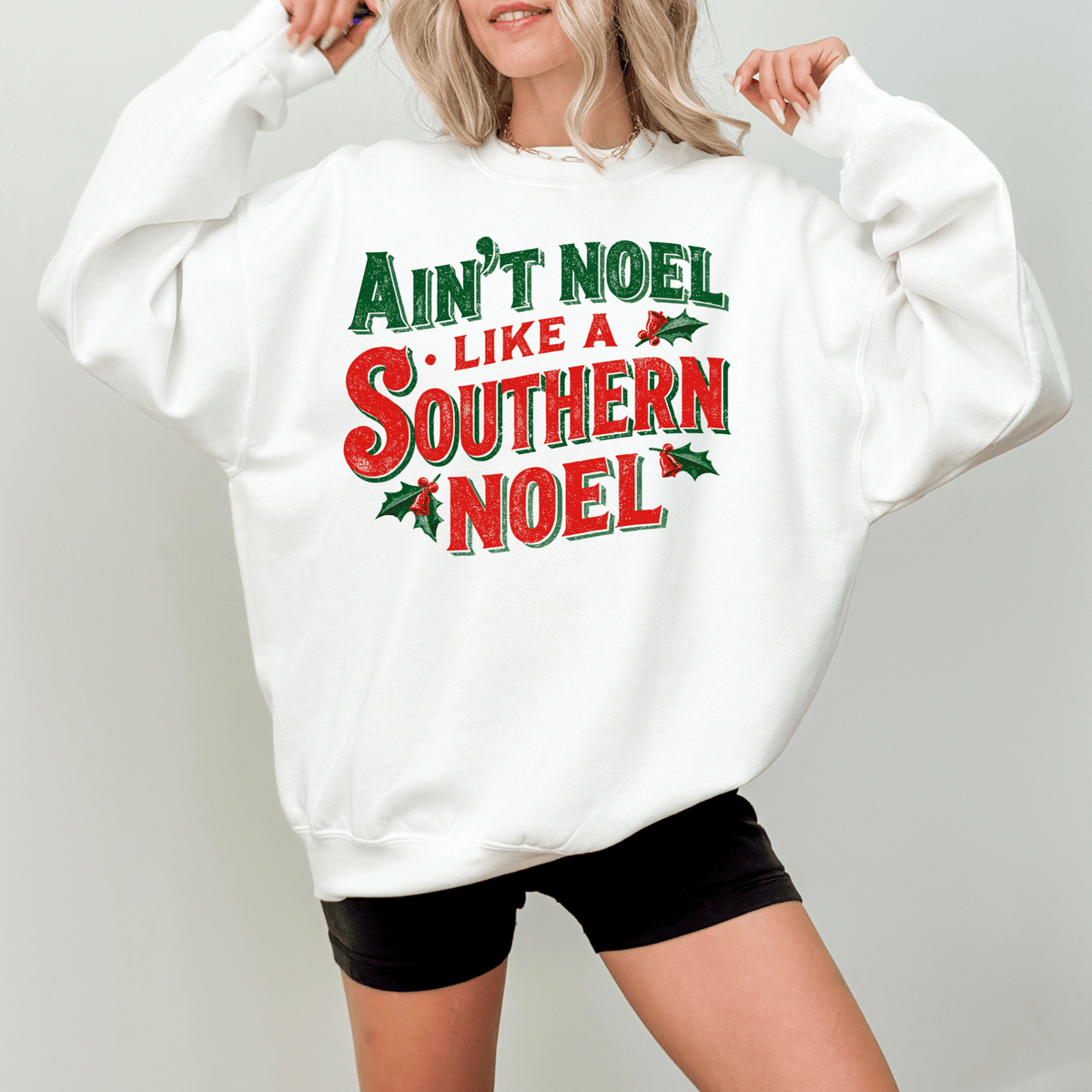 A woman wearing a white sweatshirt with a red and green text design that says "Ain't Noel Like a Southern Noel".