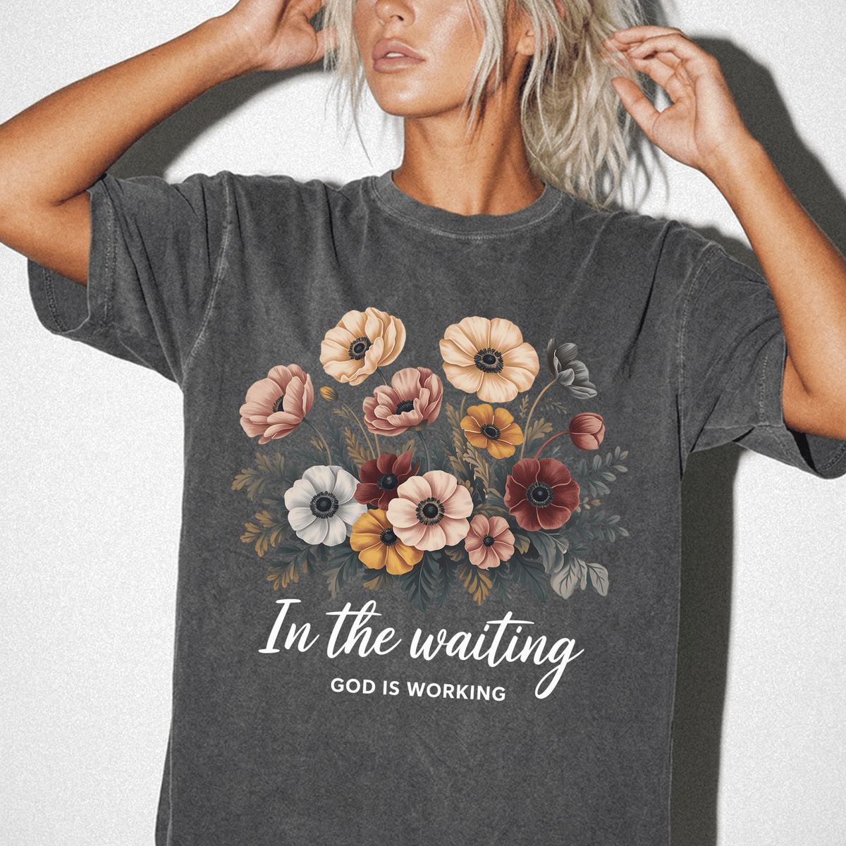 A woman wearing a gray t-shirt with a floral design and the text "In the waiting" and "God is working" printed on it.