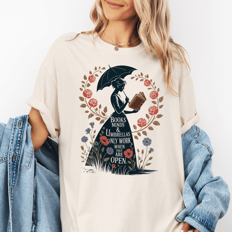 A woman wearing a white t-shirt with a graphic design of a woman holding an umbrella and reading a book, surrounded by flowers and the text "Books minds & umbrellas only work when they are open".