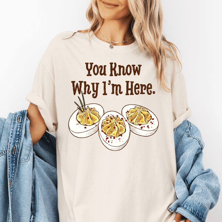 A woman wearing a white t-shirt with a graphic design of three deviled eggs and the text "You Know Why I'm Here."