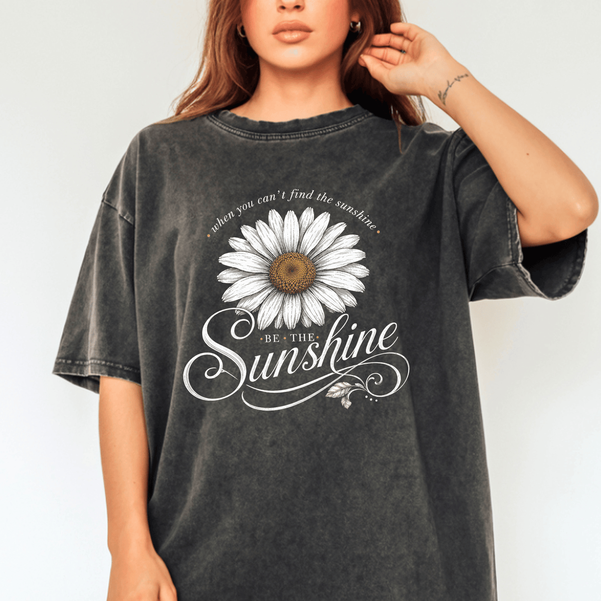 A woman wearing a gray t-shirt with a white daisy flower design and the text "Be the sunshine" printed on it.