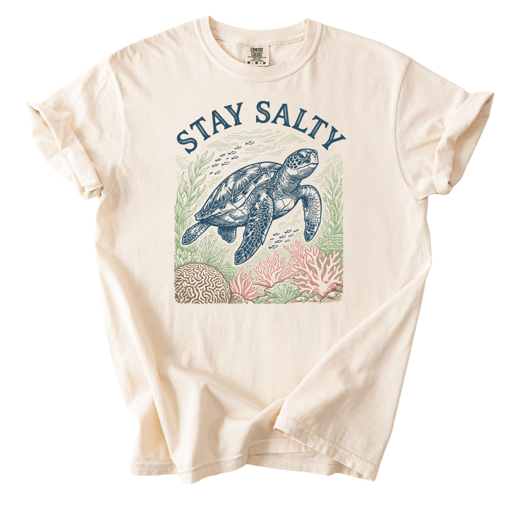 A white t-shirt with a graphic design depicting a sea turtle swimming among coral and marine life. The text above the design reads "STAY SALTY".