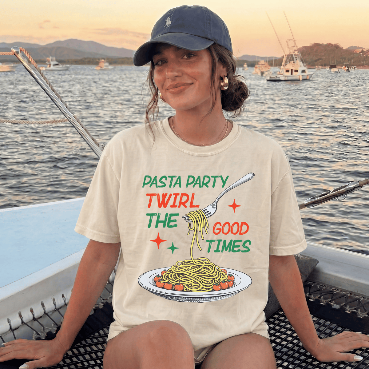 A woman wearing a t-shirt with a graphic of spaghetti and a fork, sitting on a boat with a scenic background of water and boats.