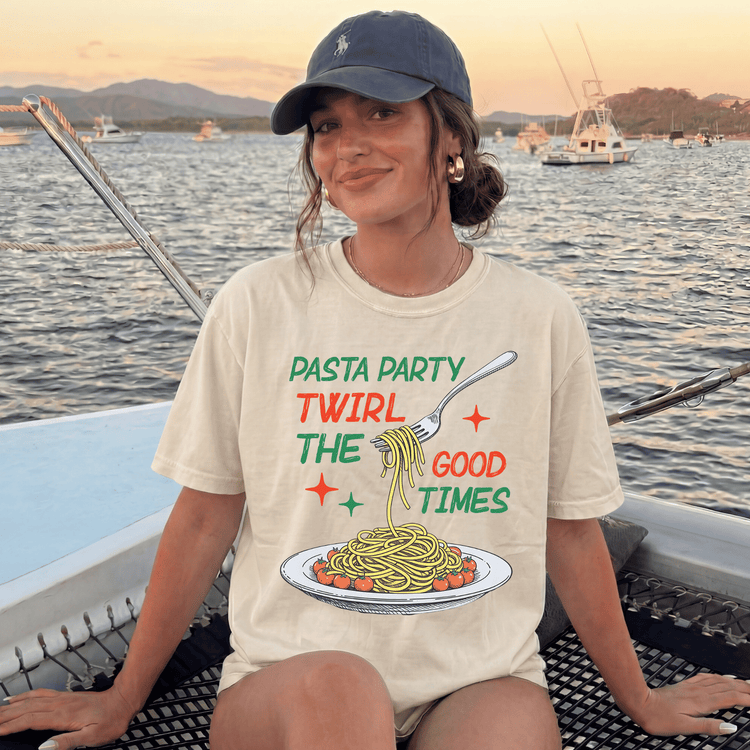 A woman wearing a t-shirt with a graphic of spaghetti and a fork, sitting on a boat with a scenic background of water and boats.