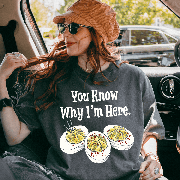 A woman wearing a hat and sunglasses is sitting in a car, with a t-shirt featuring a graphic of three deviled eggs and the text "You Know Why I'm Here."