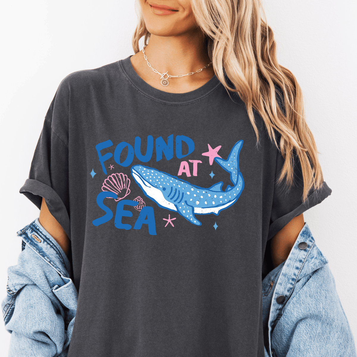 A woman wearing a gray t-shirt with a graphic design of a blue whale and seashells, along with the text "FOUND AT SEA".