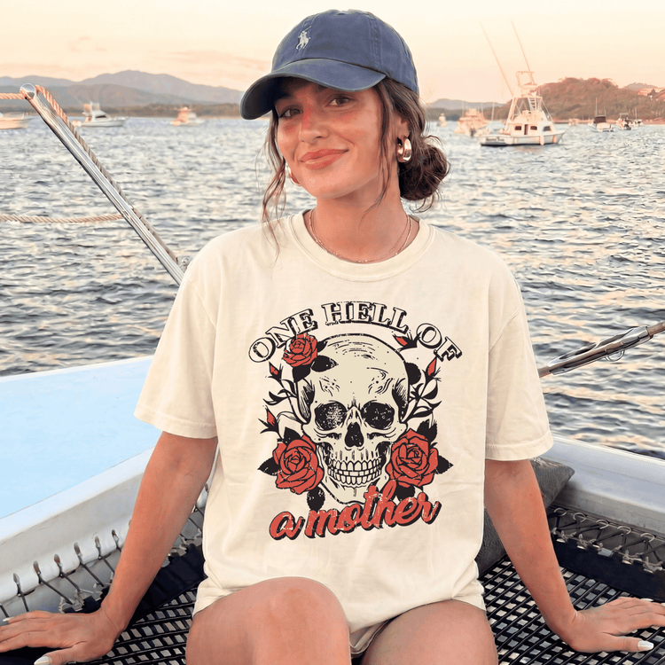 A woman wearing a white t-shirt with a skull design and the text "One Hell of a Mother" is sitting on a boat in the water.