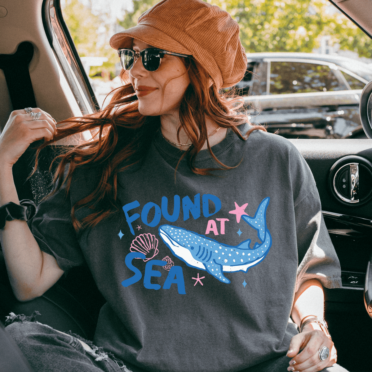 A woman wearing a hat and sunglasses is sitting in a car, looking out the window. She is wearing a gray t-shirt with a graphic design of a blue whale and seashells.