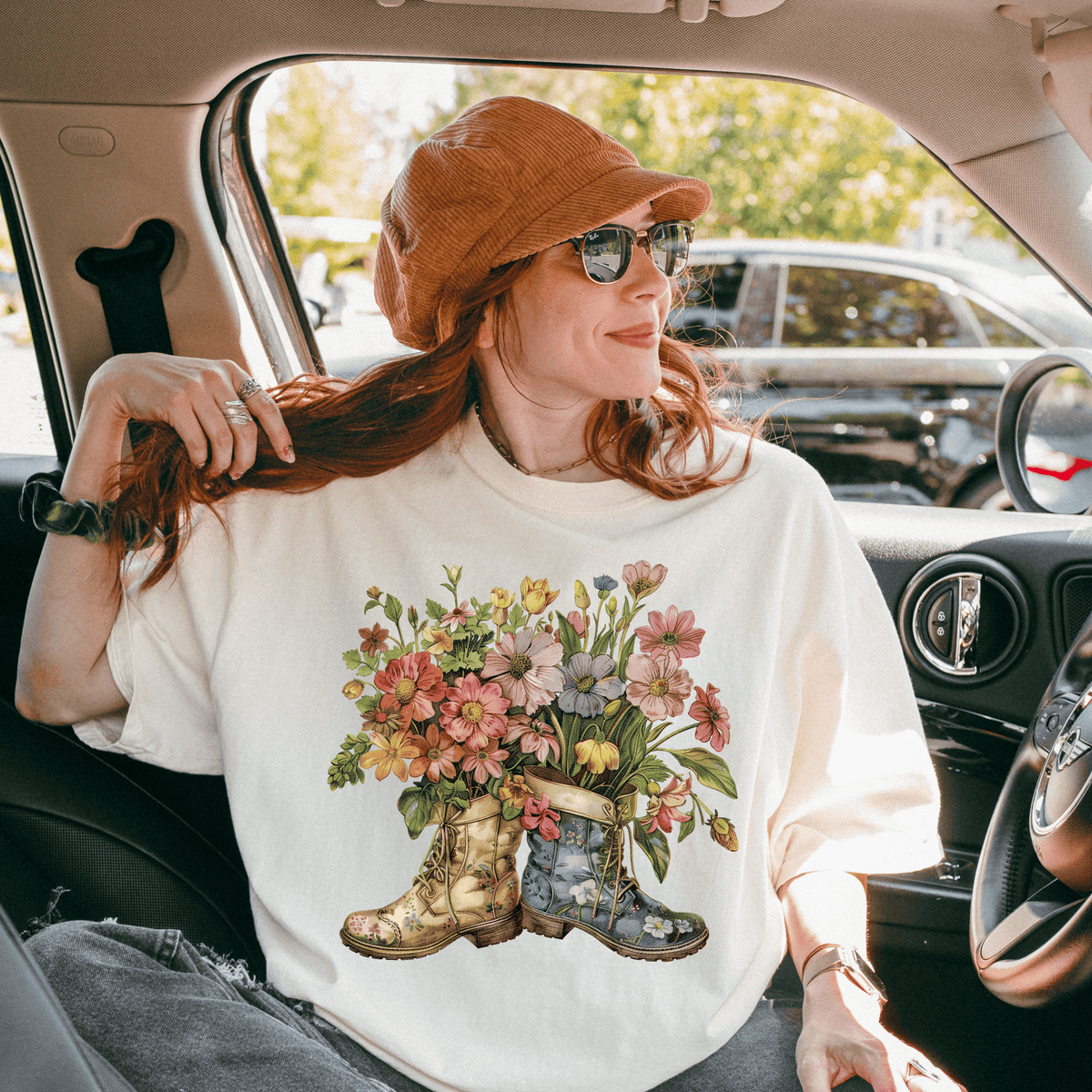 A woman with red hair is sitting in a car, wearing a white t-shirt with a floral design and a brown hat.