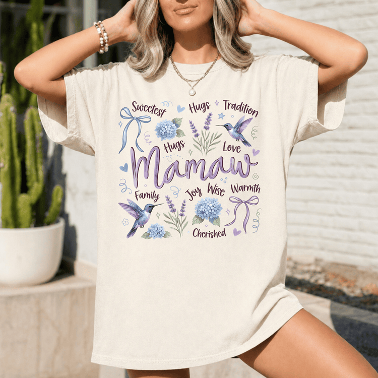 A woman wearing a white t-shirt with a graphic design featuring the word "Mamaw" surrounded by various floral and bird-related elements.