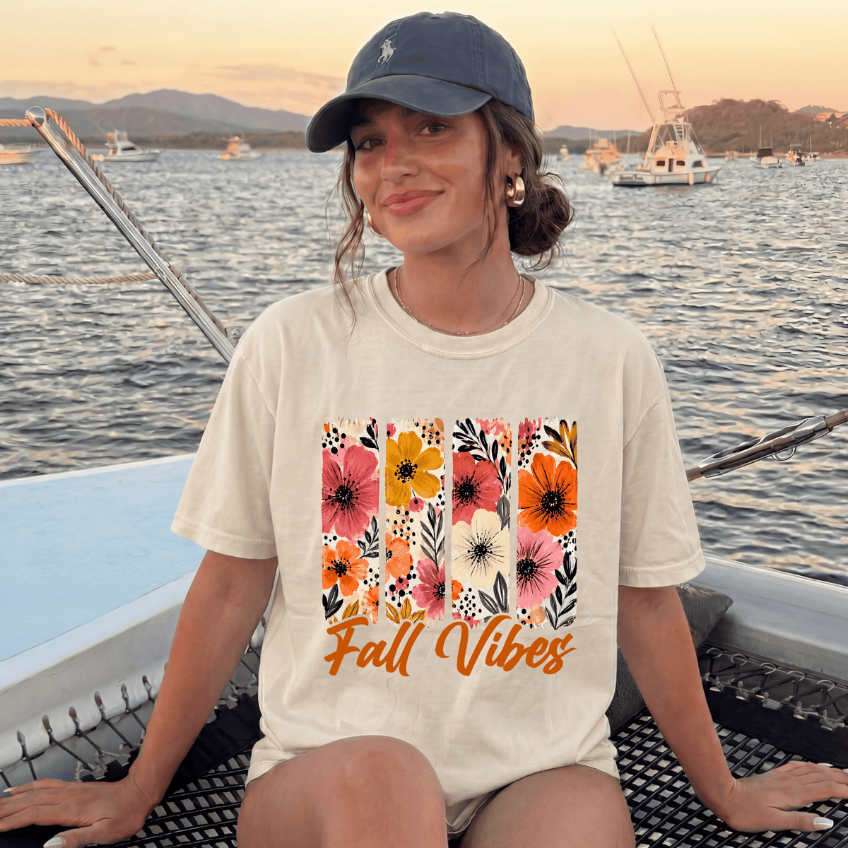 A woman wearing a white t-shirt with a floral design and the text "Fall Vibes" is sitting on a boat, smiling at the camera.