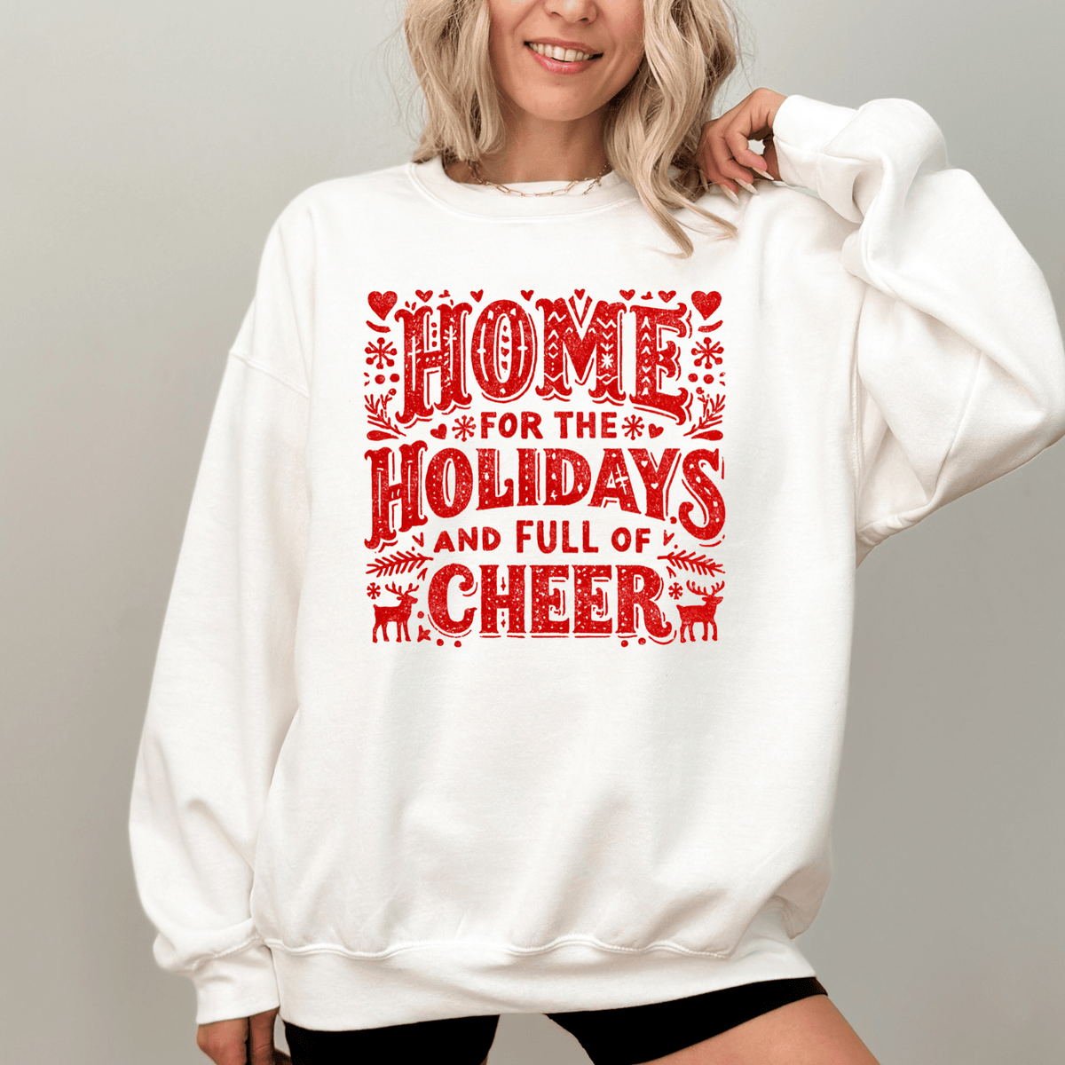 A woman wearing a white sweatshirt with a red and white graphic design that says "HOME FOR THE HOLIDAYS AND FULL OF CHEER".
