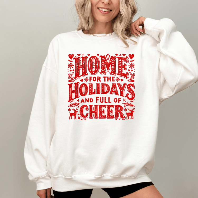 A woman wearing a white sweatshirt with a red and white graphic design that says "HOME FOR THE HOLIDAYS AND FULL OF CHEER".