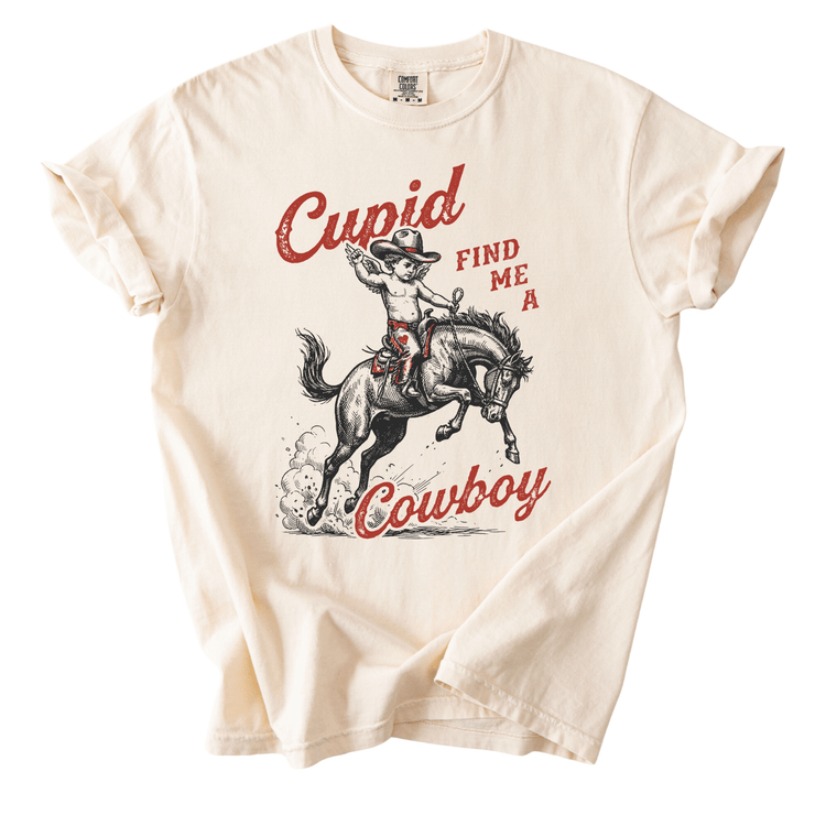 A t-shirt with a graphic design depicting a cowboy riding a horse, accompanied by the text "Cupid FIND ME A Cowboy" in a playful, humorous style.