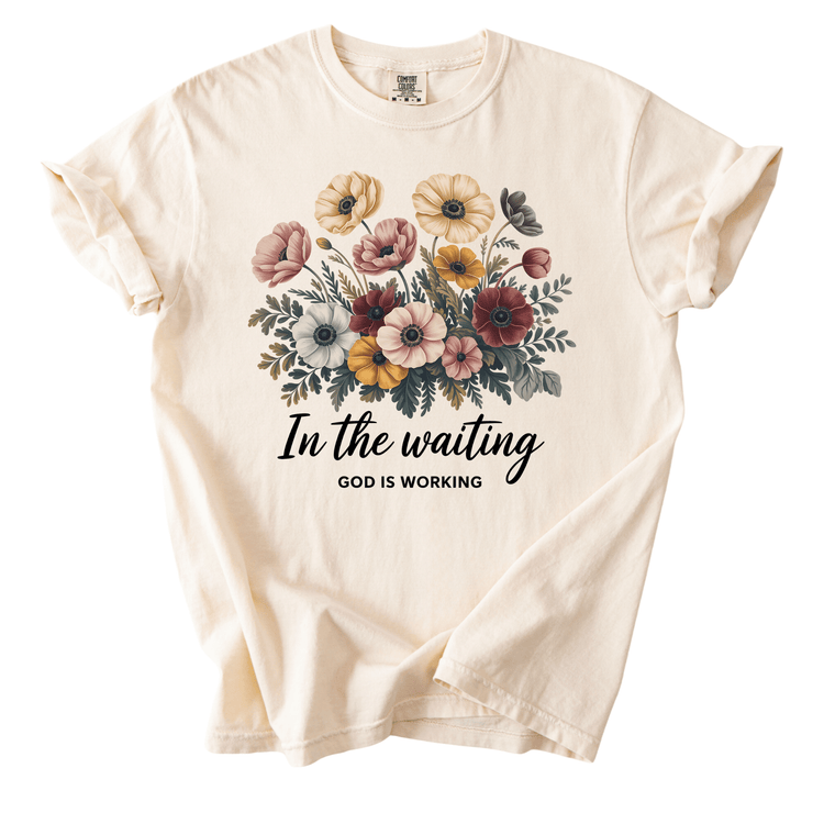 A white t-shirt with a floral design and the text "In the waiting, God is working" printed on it.