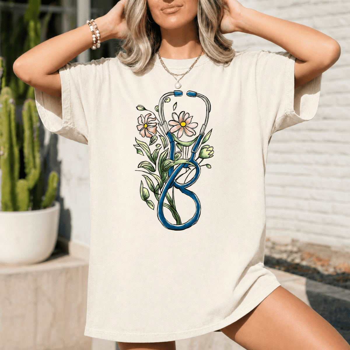 A woman wearing a white t-shirt with a graphic design of a stethoscope surrounded by flowers and leaves.