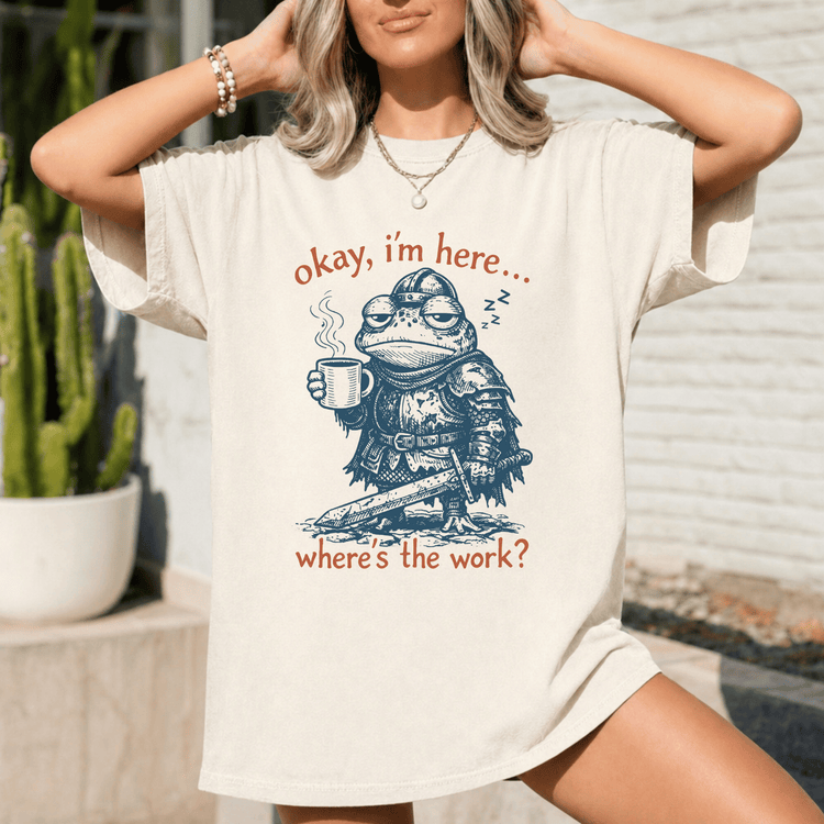 A woman wearing a white t-shirt with a graphic design of a frog holding a cup and posing with the text "okay, I'm here... where's the work?"