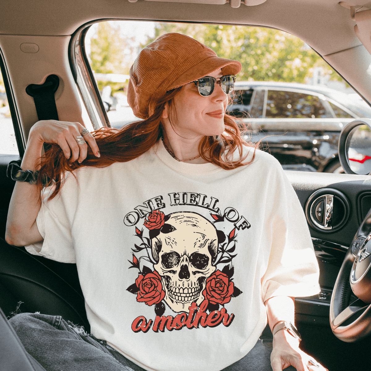 A woman wearing a white t-shirt with a skull design and the text "One Hell of a Mother" is sitting in a car.
