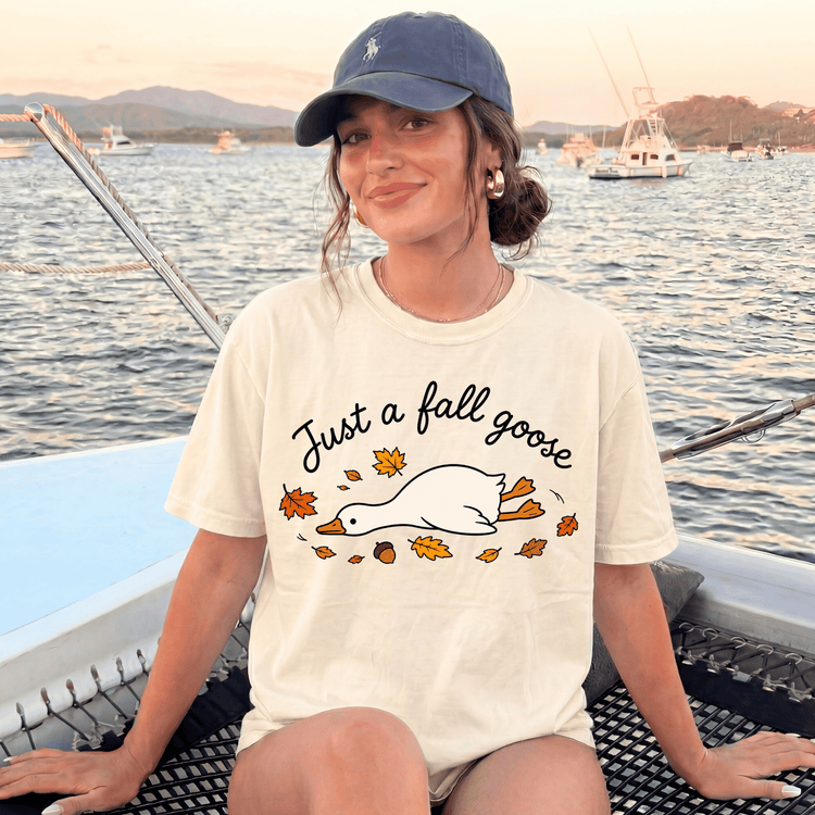 A woman wearing a white t-shirt with a graphic of a goose and autumn leaves, sitting on a boat with a scenic background of mountains and boats.