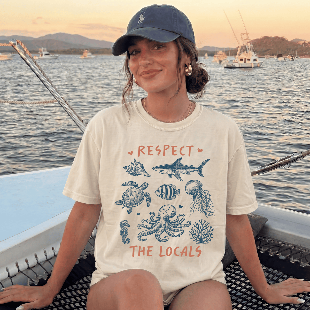 A woman wearing a white t-shirt with a graphic design of marine life and the text "RESPECT THE LOCALS" is sitting on a boat in the ocean.