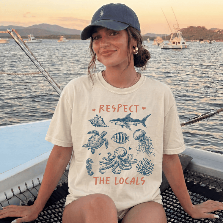 A woman wearing a white t-shirt with a graphic design of marine life and the text "RESPECT THE LOCALS" is sitting on a boat in the ocean.