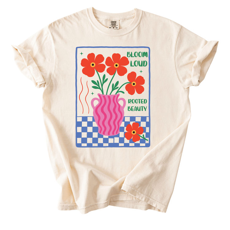 A white t-shirt with a graphic design featuring a pink vase with red flowers, a checkered background, and the text "Bloom Loud Rooted Beauty".