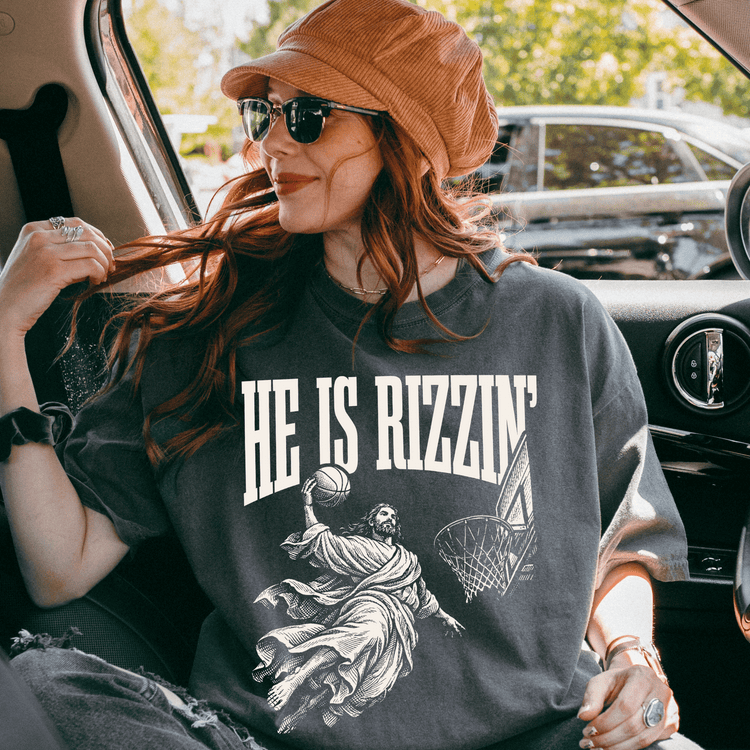 A woman wearing a hat and sunglasses is sitting in a car, looking out the window. She is wearing a gray t-shirt with a graphic design on it.