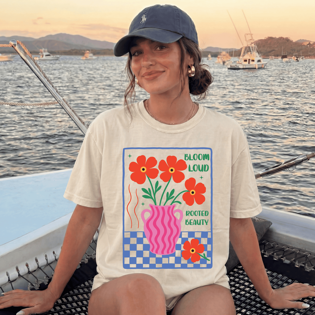 A woman wearing a white t-shirt with a graphic design of flowers and a pink vase, sitting on a boat with a scenic background of mountains and boats.