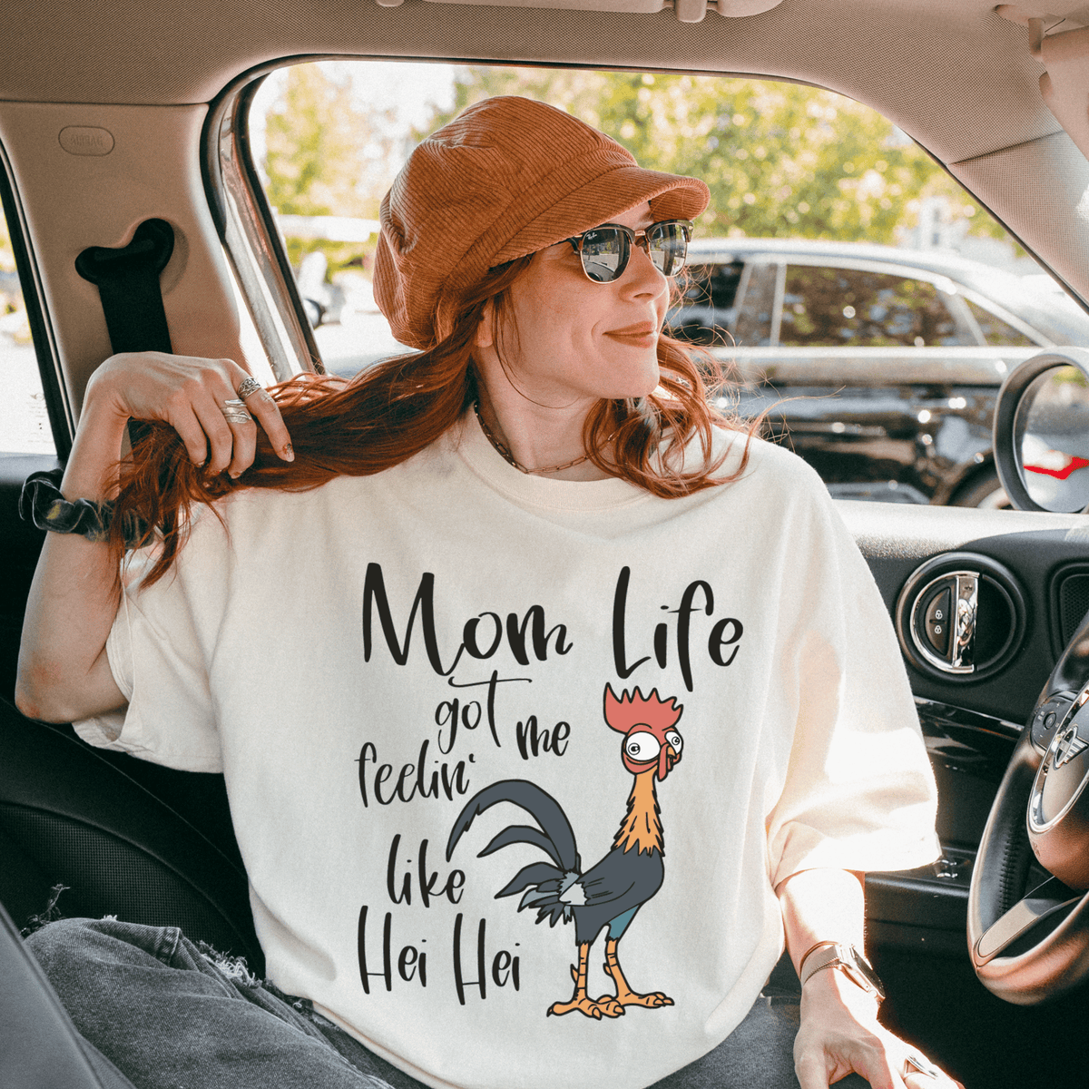 A woman wearing a white t-shirt with a graphic of a rooster and the text "Mom Life got me feelin' like Hei Hei" is sitting in a car.