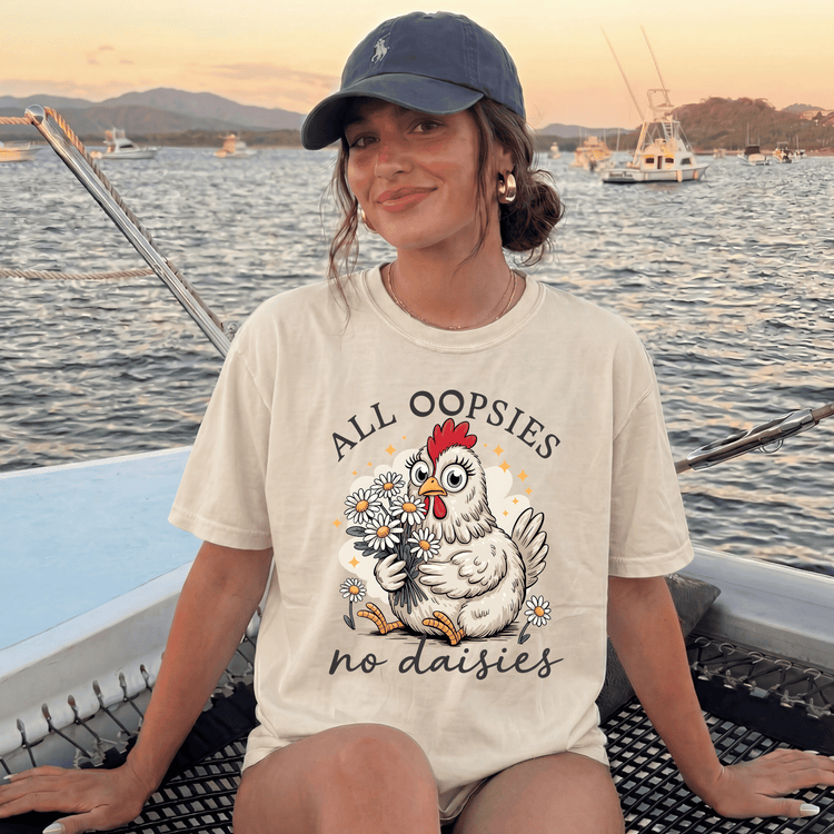 A woman wearing a white t-shirt with a graphic of a chicken holding flowers and the text "Alloopies no daisies" is sitting on a boat in the water.