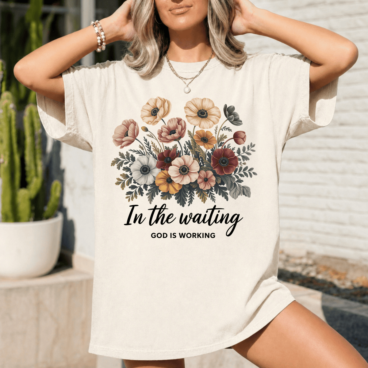 A woman wearing a white t-shirt with a floral design and the text "In the waiting, God is working" printed on it.