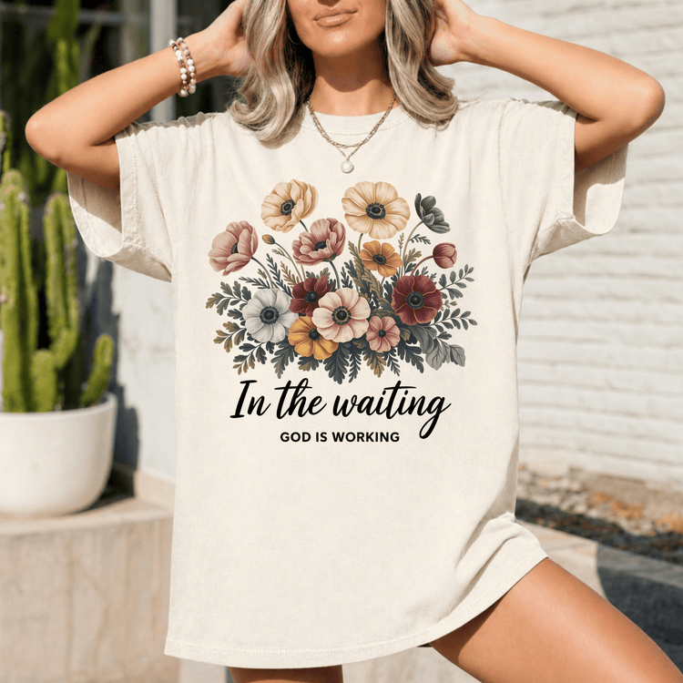A woman wearing a white t-shirt with a floral design and the text "In the waiting, God is working" printed on it.