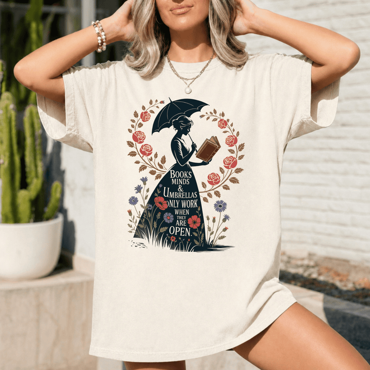 A woman wearing a white t-shirt with a graphic design of a woman holding an umbrella and reading a book, surrounded by flowers and a quote about books and minds.