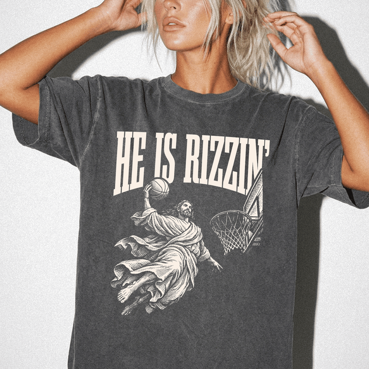 A woman wearing a black t-shirt with a graphic design depicting a basketball player in mid-air, with the text "HE IS RIZZIN" printed on the shirt.