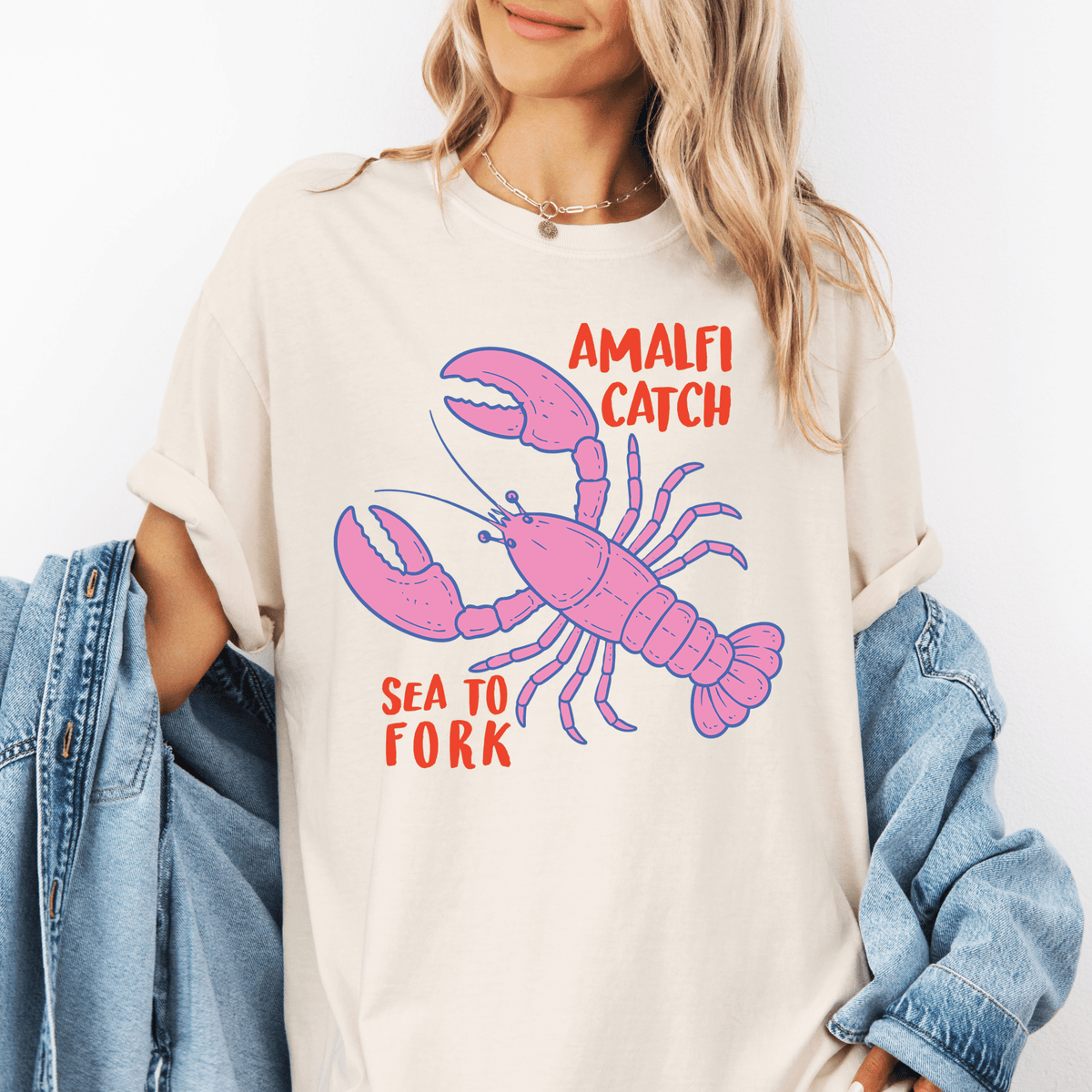 A woman wearing a white t-shirt with a graphic of a pink lobster and the text "Amalfi Catch Sea to Fork" printed on it.