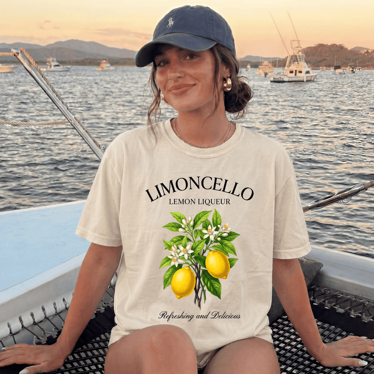 A woman wearing a cap and a white t-shirt with a lemon graphic is sitting on a boat, smiling at the camera.