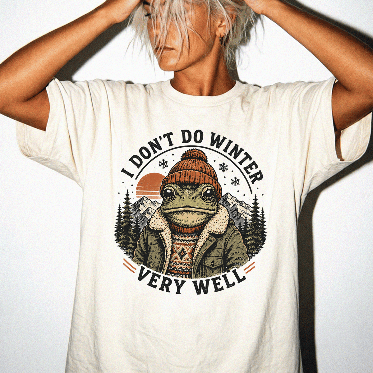 A person wearing a white t-shirt with a graphic design of a frog wearing winter clothing and a hat, with mountains in the background.