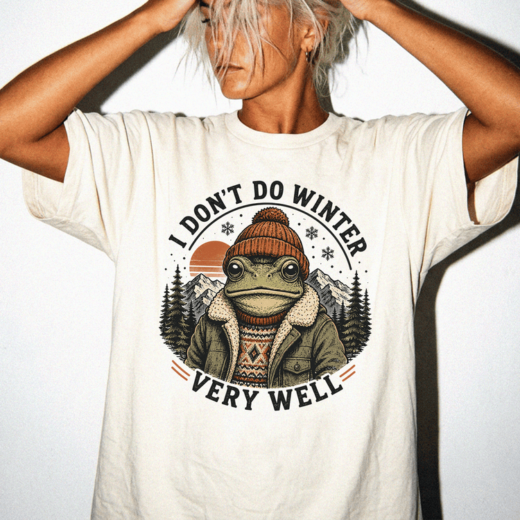 A person wearing a white t-shirt with a graphic design of a frog wearing winter clothing and a hat, with mountains in the background.
