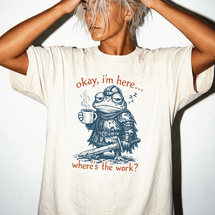 A person wearing a white t-shirt with a graphic design depicting a frog character holding a cup and posing with the text "okay, I'm here... where's the work?"