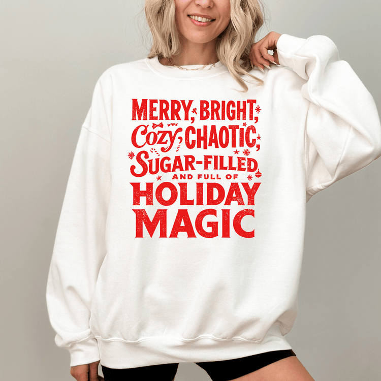 A woman wearing a white sweatshirt with a red text design that says "MERRY, BRIGHT, COZY, CHAOTIC, SUGAR-FILLED, AND FULL OF HOLIDAY MAGIC".