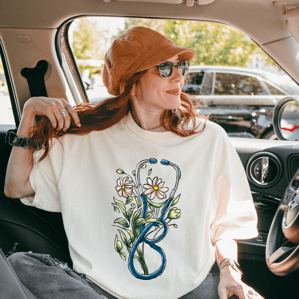 A woman with red hair is sitting in the passenger seat of a car, wearing a white t-shirt with a stethoscope design and a brown hat.