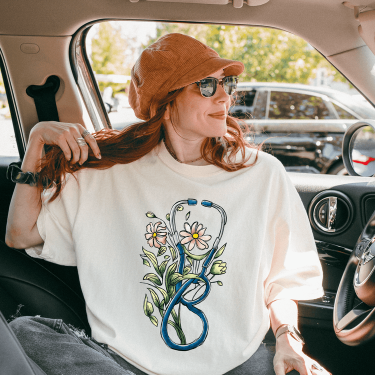 A woman with red hair is sitting in the passenger seat of a car, wearing a white t-shirt with a stethoscope design and a brown hat.