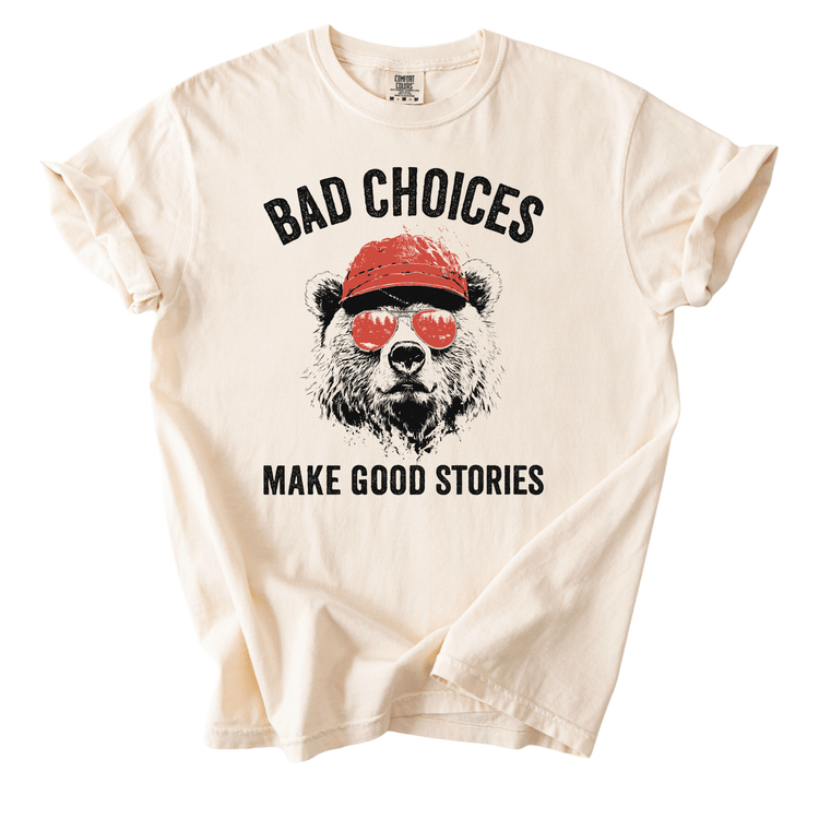 A white t-shirt with a graphic design of a bear wearing sunglasses and a red hat, along with the text "Bad choices make good stories" printed on it.