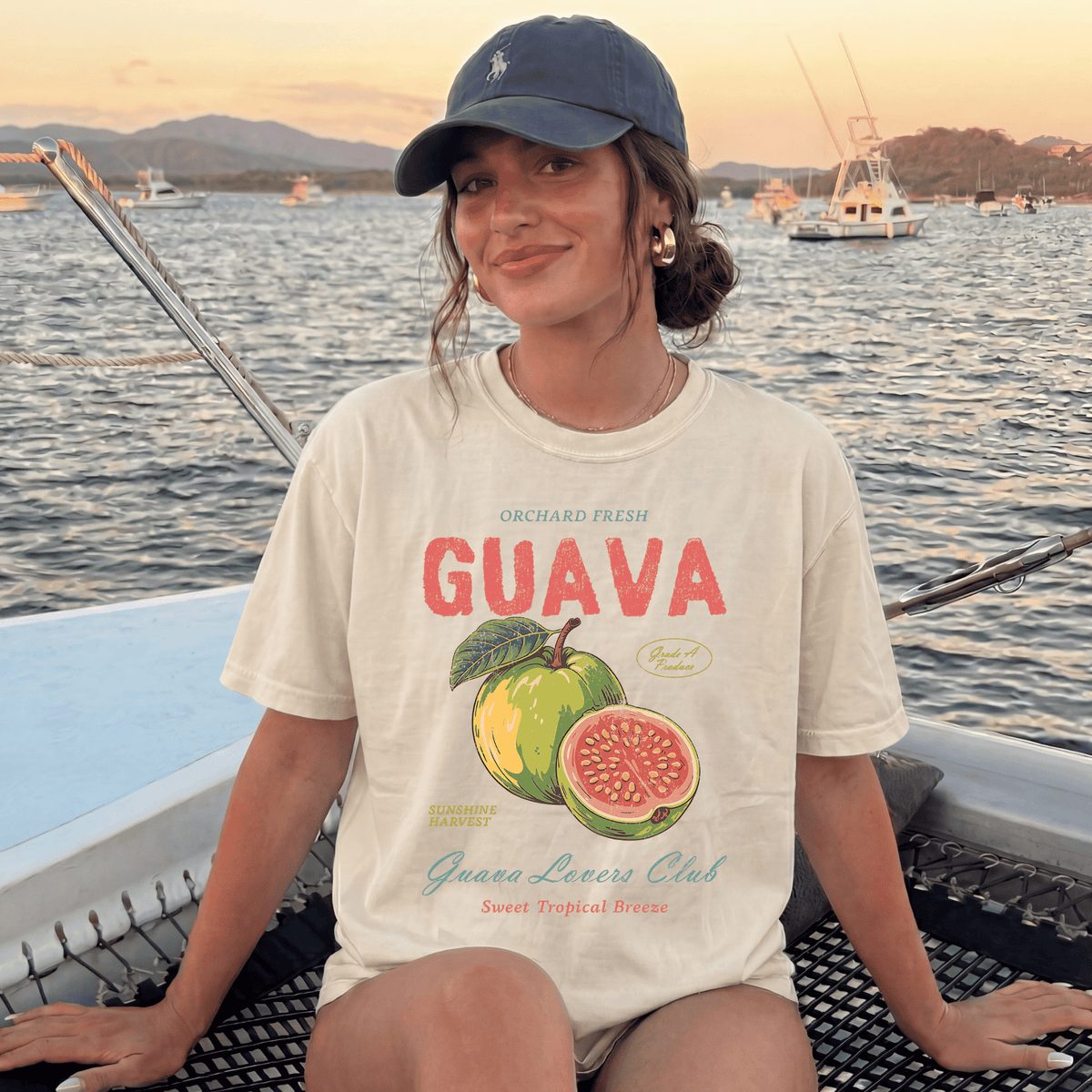 A woman wearing a white t-shirt with a graphic of a guava and text, sitting on a boat with a scenic background of water and boats.