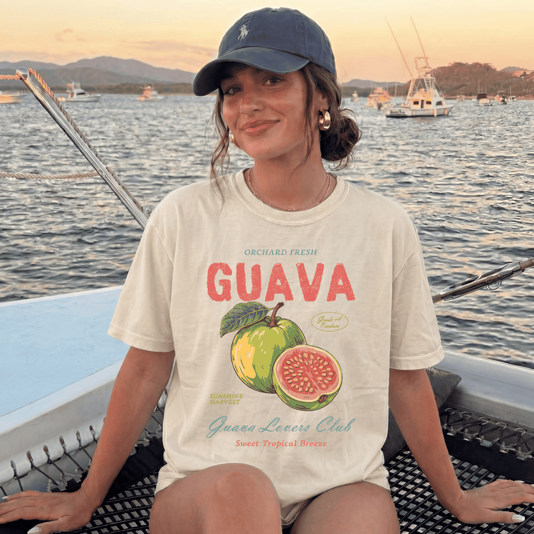 A woman wearing a white t-shirt with a graphic of a guava and text, sitting on a boat with a scenic background of water and boats.